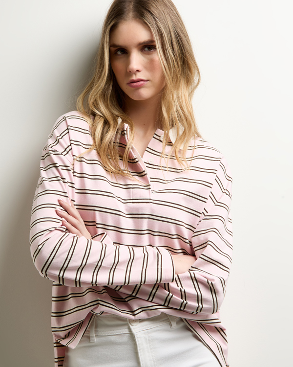 Young woman with long hair wears a pink and white striped shirt and white pants, standing against a white wall with arms crossed and a confident expression.