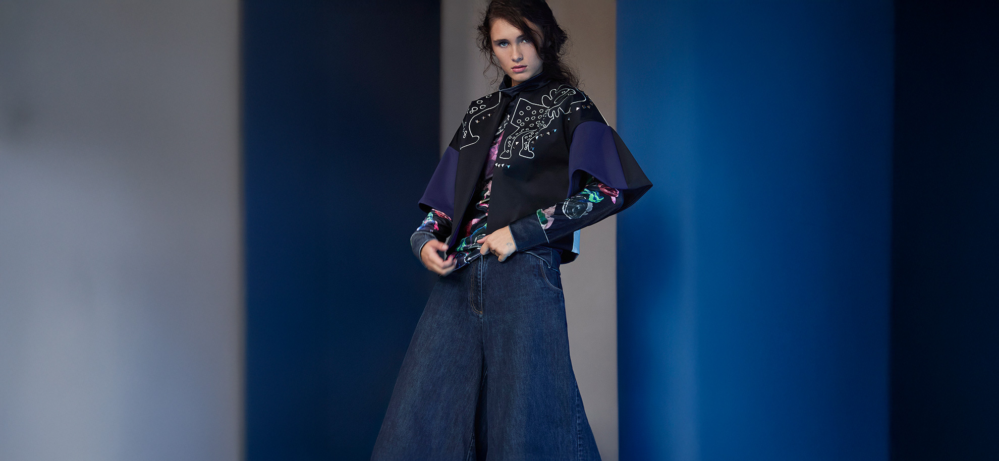 A person wearing a stylish outfit with a graphic, animal-patterned cape and floral sleeves stands against a blue and beige background, exuding a confident vibe.