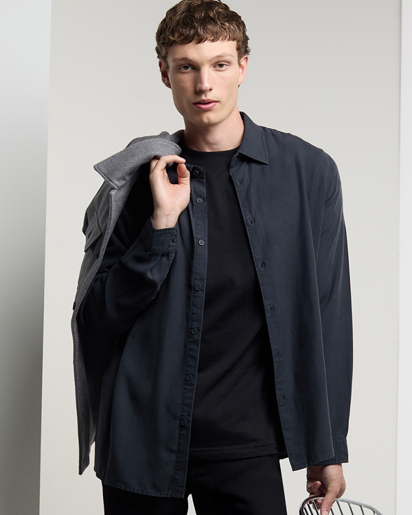 Young man with short hair, wearing a dark shirt over a black t-shirt, carries a gray jacket over his shoulder. He stands against a neutral backdrop.