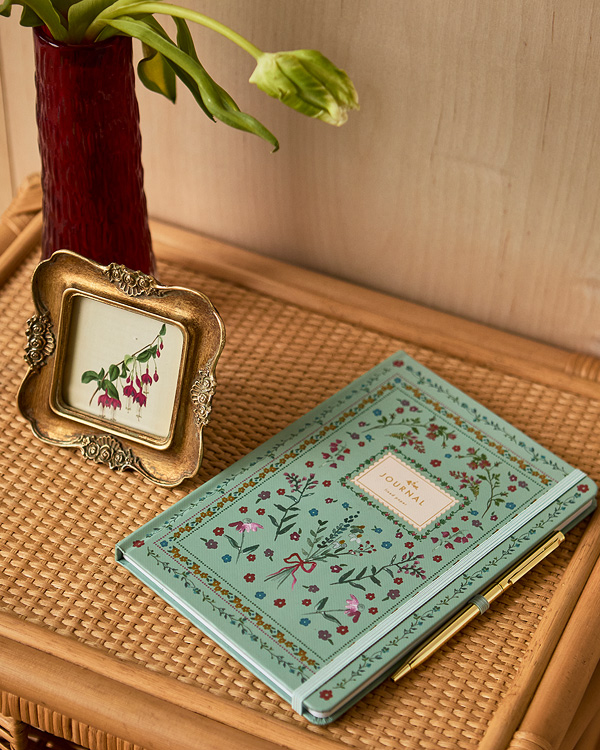 A teal journal with floral designs lies on a wicker surface next to a red vase with drooping flowers and an ornate gold-framed botanical print.