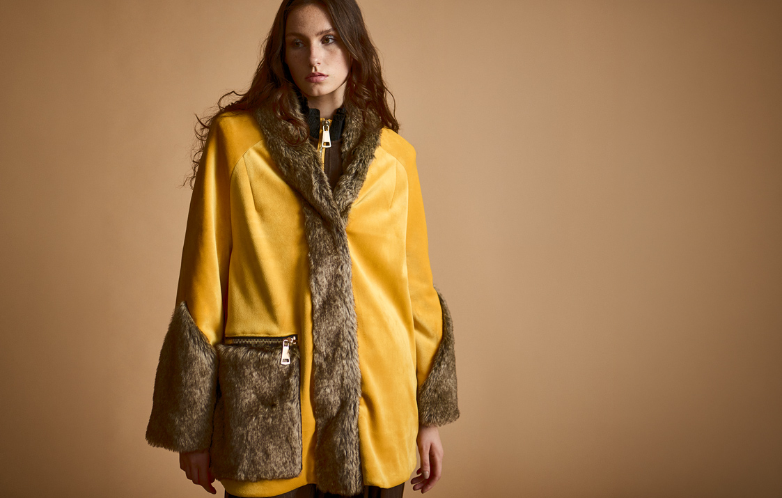 A woman poses against a tan backdrop wearing a bright yellow velvet coat accented with brown faux-fur trim on the collar, cuffs, and front zipper pocket.