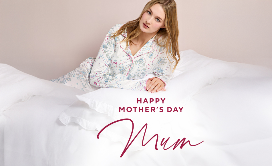 Woman in floral pajamas reclines on a white bed, looking serene. Text below reads "Happy Mother's Day Mum" in elegant, red script.